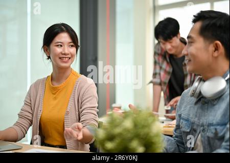 An attractive and cheerful young Asian female worker in a startup team is talking and sharing her ideas with a male coworker in the office. Stock Photo