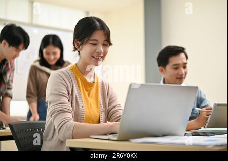 Close-up image of a beautiful and smart young Asian female worker in a startup team is working on her tasks on hera laptop while her colleagues are in Stock Photo