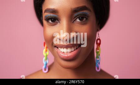 Color makeup. Happy face. Summer beauty. Cheerful smiling woman with full lips blue eye shadow long eyelashes rainbow earrings on pink free space. Stock Photo