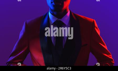 Gentleman fashion. Dandy style. Party look. Neon light unrecognizable confident man in red elegant tuxedo suit on dark fluorescent purple color backgr Stock Photo