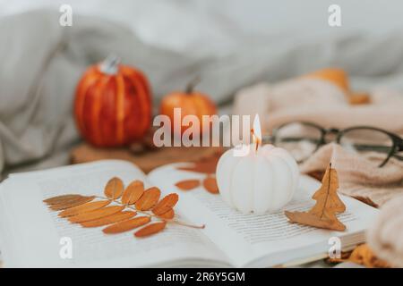 White burning candle shape of pumpkin on open book, pumpkins decor, eyeglasses and autumn leaves on bed in cozy domestic atmosphere Stock Photo