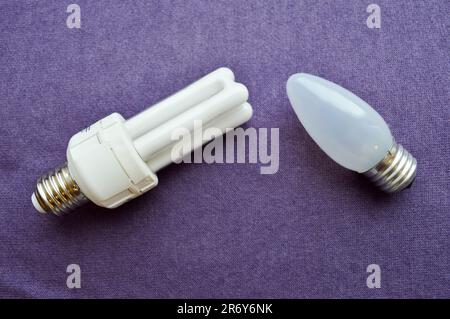 Two light bulbs. White energy-saving fluorescent bulb with four tubes, with a silver base and an oblong incandescent bulb with gray glass on a purple Stock Photo