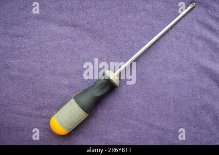 Screwdriver with a rubberized yellow-gray handle and a cross-shaped slot on the background of a purple cloth. Manual metalwork tools. Stock Photo