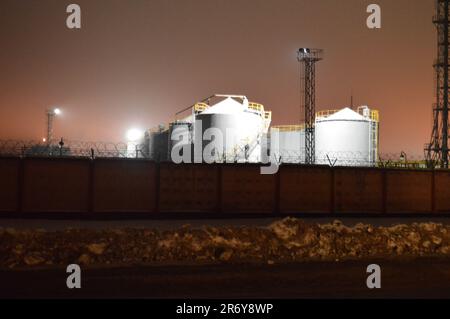 Night photo of a tank farm of a petroleum refinery, a petrochemical plant through a fence with a barbed wire. Containers with fuel. Stock Photo