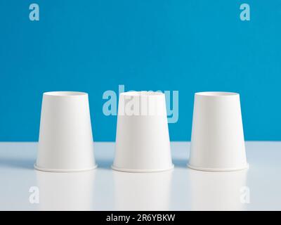 Three upside down flipped white paper cups on blue background. Stock Photo