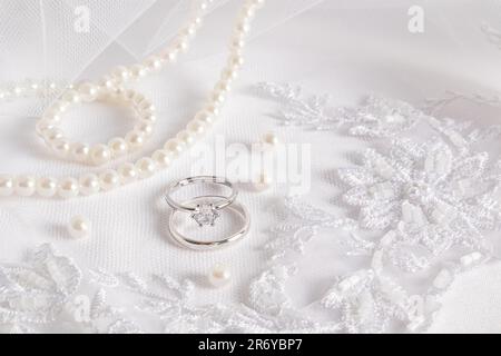 Gentle wedding background. Two white gold engagement rings with a diamond on a white embroidered veil and a pearl necklace. space for text Stock Photo