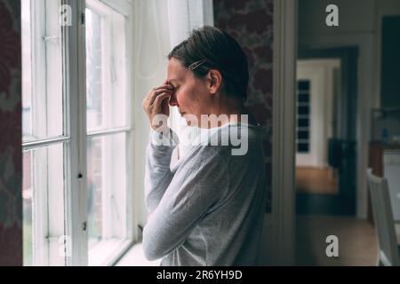Mid-adult caucasian woman with severe headache standing by the living room window in morning and pressing the forehead with fingers, selective focus Stock Photo