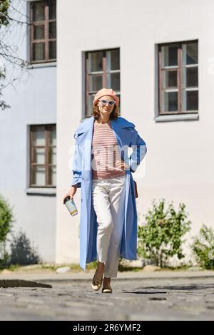 Attractive adult woman in a fashionable, stylish coat walks on a pavement holding a cardboard cup for coffee in her hand Stock Photo