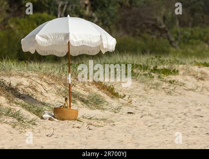 A white beach umbrella sits in the sand on a sunny day with trees in the background Stock Photo