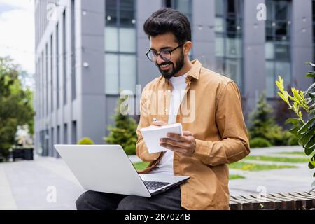 Young Indian man studying online sitting on bench outside university campus, student with laptop taking notes on seminar, smiling happy with remote learning. Stock Photo