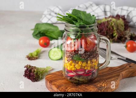 Healthy salad with quinoa, cucumbers, corn, cherry tomato, radish, spinach, lettuce and olive oil in a Mason jar on gray background Stock Photo