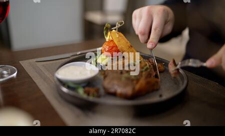 A man cuts a cooked steak from medium-sized ribs. Delicious steak with vegetables on the table. A man in a restaurant eats, cuts meat with a knife. Stock Photo