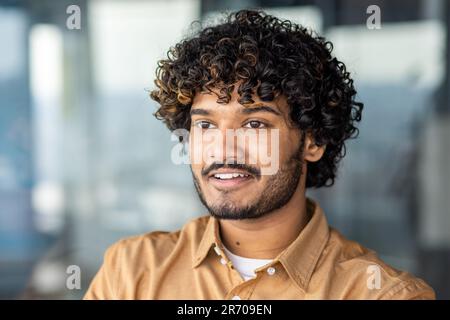 Close up portrait of young Hindu programmer, man in shirt smiling and looking out window, successful businessman inside office at workplace, satisfied financier accountant with curly hair. Stock Photo