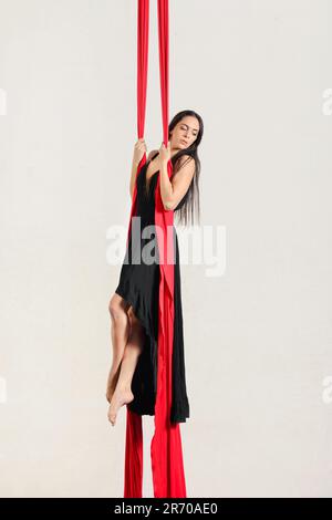Side view of gracious young barefooted female performer with long dark hair in black dress hanging on red aerial silks and looking over shoulder again Stock Photo
