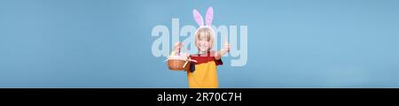 Happy boy with bunny ears holding basket full of Easter eggs and showing thumbs up on light blue background. Banner design Stock Photo