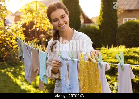 Smiling woman hanging baby clothes with clothespins on washing line for drying in backyard Stock Photo
