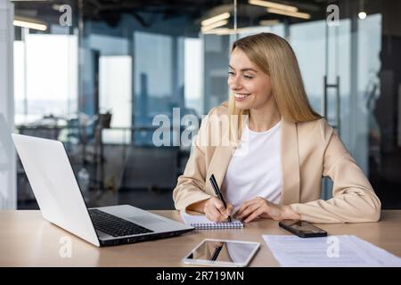 Young smiling and successful businesswoman, manager works in the office with a laptop online, talks on a video call, listens and watches business training, makes notes in a notebook. Stock Photo