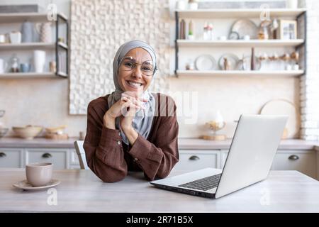 Portrait of a young beautiful Muslim female student in a hijab studying remotely on a laptop, smiling and looking at the camera. Stock Photo