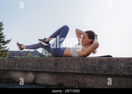 Side view of young sporty woman doing bicycle crunches on gym mat in the city. Stock Photo