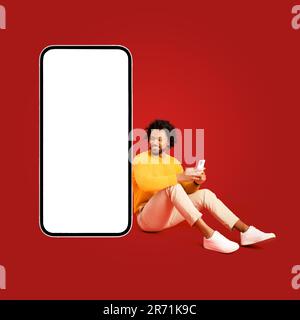 Handsome African-American curly 30s man using smartphone sitting near huge mobile phone with empty screen isolated on red, charismatic guy recommends and advertising mobile app, copy space Stock Photo