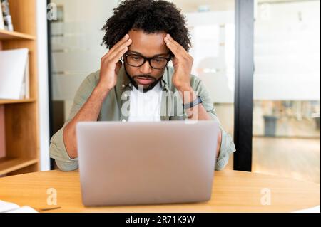 Worried african-american male employee colleague staring at laptop screen sitting on the workplace, pensive concerned businessman has problem with project, lack of new ideas Stock Photo