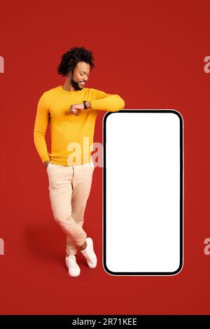 Best deal, promotion. Smiling positive african-american man leaning on huge smartphone with empty blank screen, looking at it, freelancer guy recommending new app or website, mockup Stock Photo