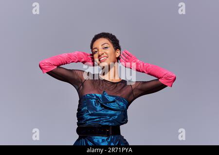 Joyful and short haired african american woman with bold makeup wearing pink gloves and cocktail dress while standing and posing isolated on grey, mod Stock Photo