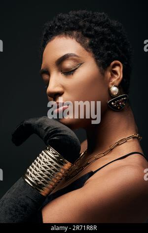 Portrait of fashionable african american woman in dress, glove and golden accessories touching chin while standing isolated on black, high fashion and Stock Photo