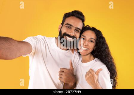Cheerful Couple Making Selfie Gesturing Thumbs Up Over Yellow Background Stock Photo