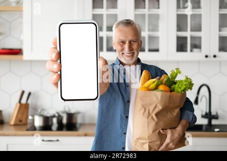 Grocery Delivery App. Smiling Senior Man Showing Blank Smartphone And Holding Groceries Stock Photo