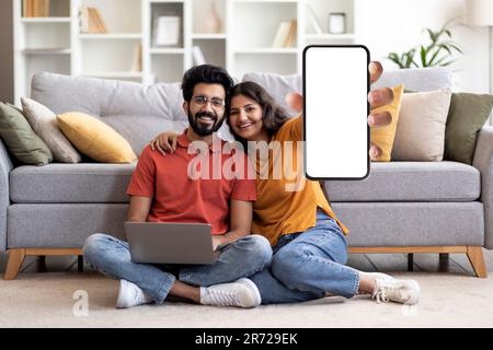 Happy Young Indian Couple Showing Blank Smartphone And Using Laptop At Home Stock Photo