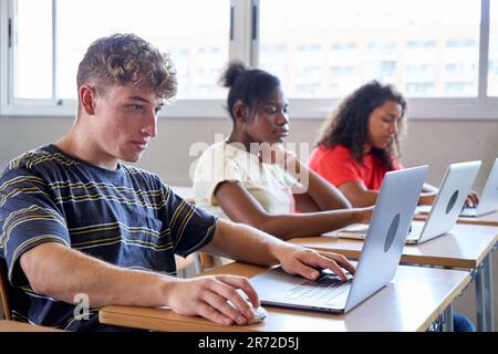 Side view of teenage students in a classroom studying with technological equipment, using laptops. Stock Photo