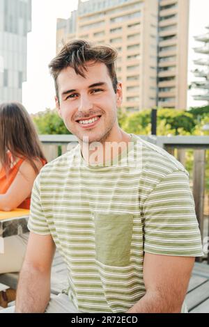 Vertical portrait of a positive and smiling young male student looking at camera sitting outside at the University Campus. Front view of handsome man laughing with cheerful and friendly attitude. High quality photo Stock Photo