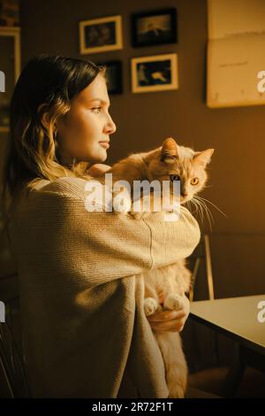 The young woman is dressed in home clothes and embracing a ginger cat. The warmth and affection that comes with caring for a cat, highlighting the uni Stock Photo