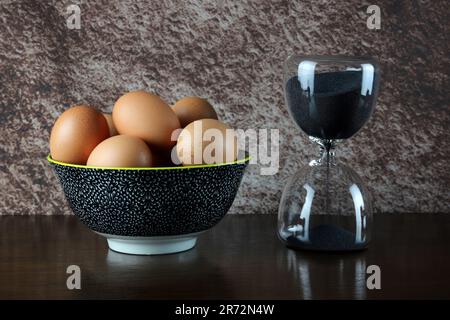 Sand glass egg timer with bowl of fresh eggs on a varnished wooden table top with a mottled background Stock Photo