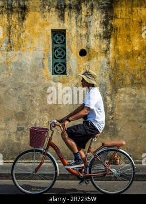 Hoian, Vietnam, March 6th, 2023. A man with hat cycling pass in front of an old building with yellow wall and small green window frame in a sunny day. Stock Photo