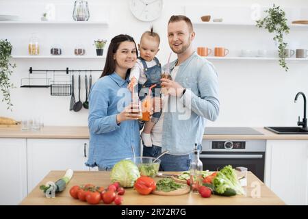 Cheerful family dressed in denim clothing smiling at camera while preparing healthy meal on table full of vegetables. Happy spouses and small toddler having fun together while making salad in kitchen. Stock Photo