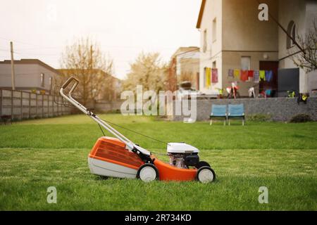 Modern orange-grey electric lawn mower on bright lush green lawn at residential backyard of house. Gardening work tools. Rotary lawn mower machine cut Stock Photo