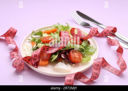 Plate with fresh vegetable salad and measuring tape on violet background, closeup. Healthy diet concept Stock Photo