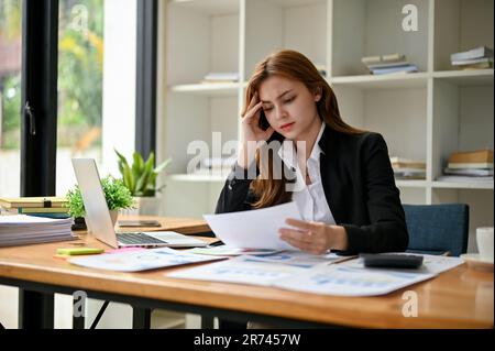 A stressed and tired Asian businesswoman focuses on reading and analyzing financial reports at her desk in her office. Stock Photo