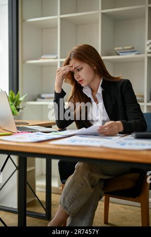 A portrait of a stressed and worried young Asian businesswoman focuses on analyzing business financial data on her laptop and working on financial rep Stock Photo