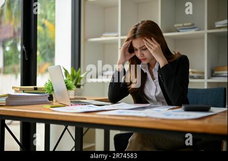 A stressed millennial Asian businesswoman focuses on working on her business financial data reports at her desk. Stock Photo