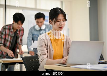A focused young Asian female marketing assistant is working on her work on her laptop at her desk while her coworkers are working in the room. Stock Photo