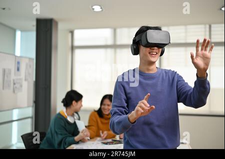 A cheerful and excited young Asian male developer tested a new VR game on a VR headset in the meeting with his team. Virtual reality, tech company, bu Stock Photo