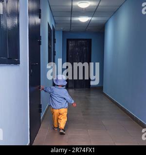 The toddler baby walks in the entrance hall on the floor near the apartment doors. A child runs in the hallway of an apartment building. Kid aged abou Stock Photo