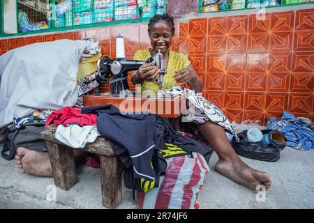 Toliara, Madagascar - May 01, 2019: Unknown older Malagasy woman sitting on ground at street next to market, mending clothes with hand crank sewing ma Stock Photo