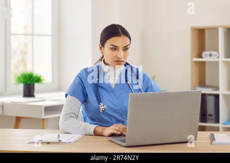 Portrait of a young female doctor in blue medical uniform working in medical office on laptop. Stock Photo