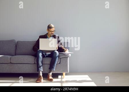 Businesslike, handsome, brutal man is sitting on the couch with laptop and making notes in notebook. Stock Photo