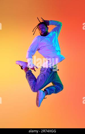 Young bearded man with dreads in casual clothes dancing breakdance against gradient orange yellow studio background in neon light Stock Photo