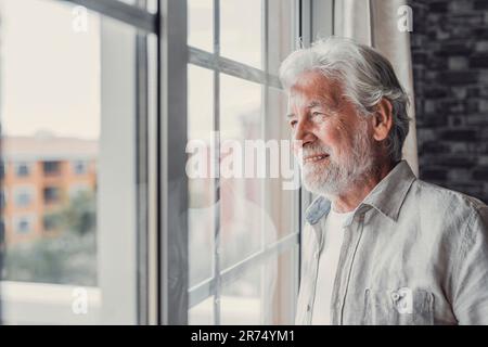 Happy thoughtful older 70s man looking out of window away with hope, thinking of good health, retirement, insurance benefits, dreaming of future. Elderly pensioner waiting meeting with family Stock Photo